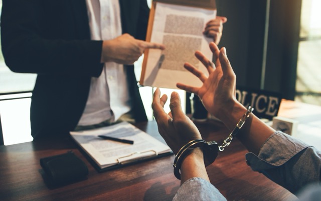 Person in handcuffs engaging with a lawyer, discussing legal documents on a table, emphasizing the importance of legal defense in criminal accusations.