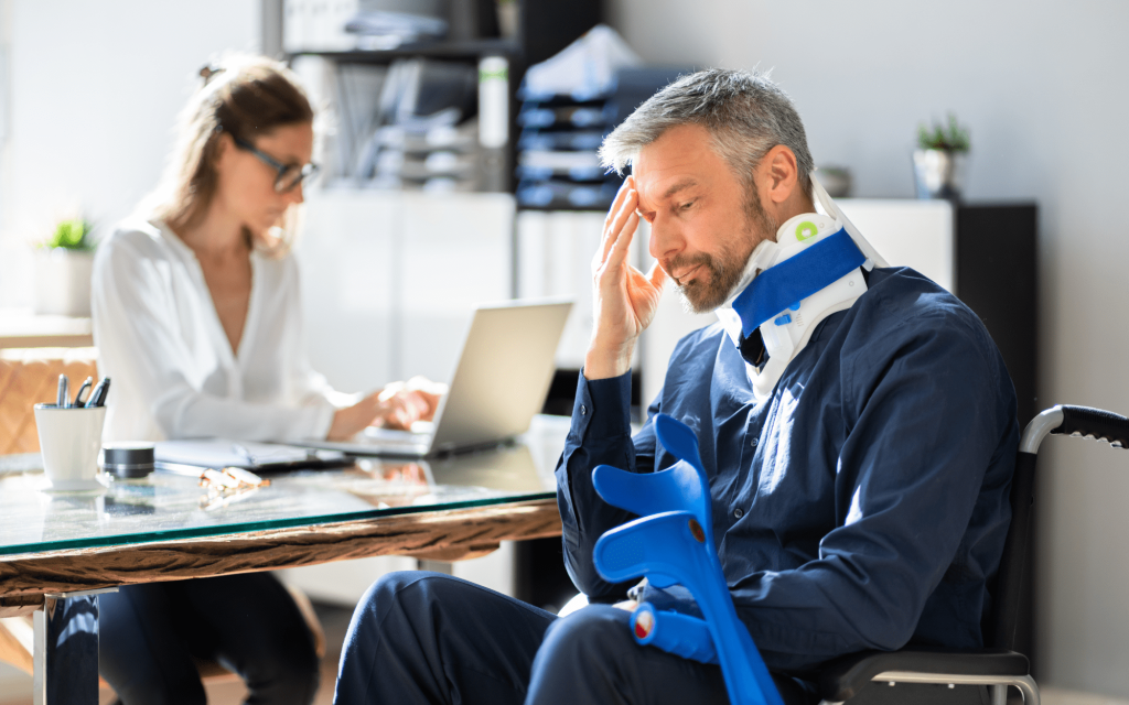 Man in a neck brace sitting in a wheelchair, looking stressed, while a woman in a white shirt works on a laptop in a law office setting, symbolizing legal support for Social Security Disability claims.