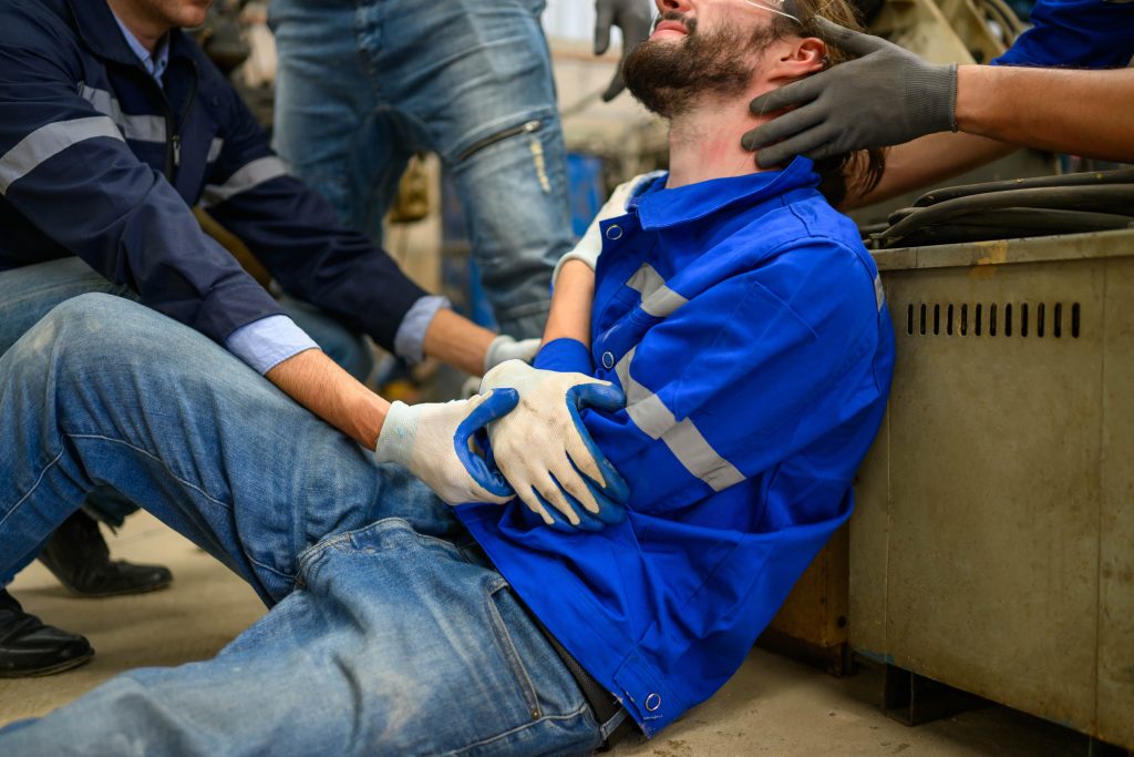 Injured factory worker receiving assistance, wearing blue work uniform, surrounded by colleagues in a hazardous industrial environment.