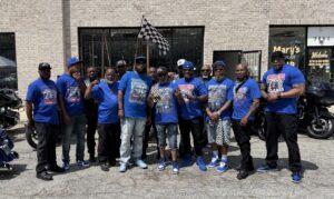 The members of the Atlanta chapter of Undisputed MC, a motorcycle enthusiasts club. The group consists of Black middle-aged men wearing jeans and posing for a photo. They wear blue t-shirts with the MC Undisputed logo on them, and their motorbikes stand in the background.
